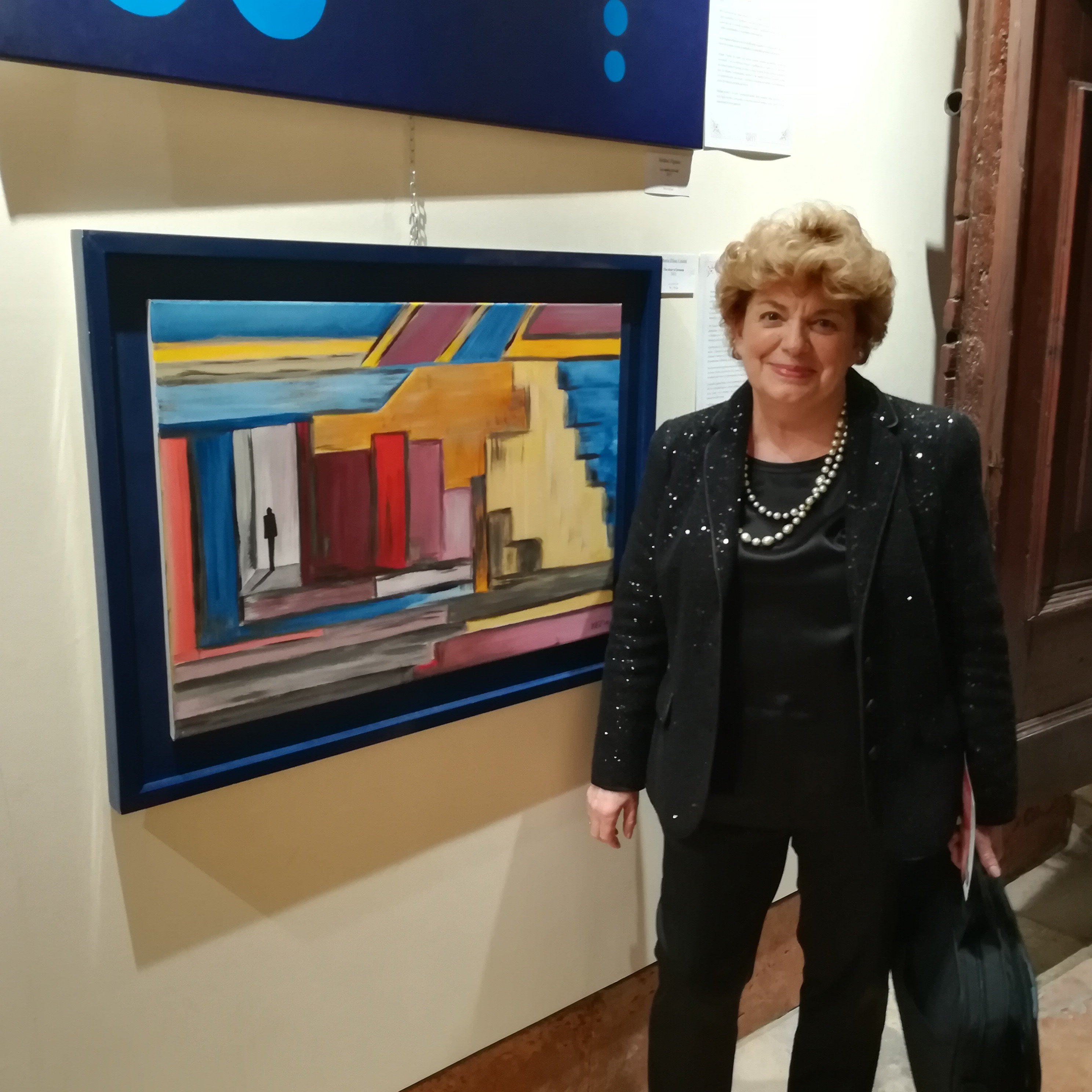 A woman standing beside a framed abstract painting in a gallery, shown in a medium view under indoor lighting.