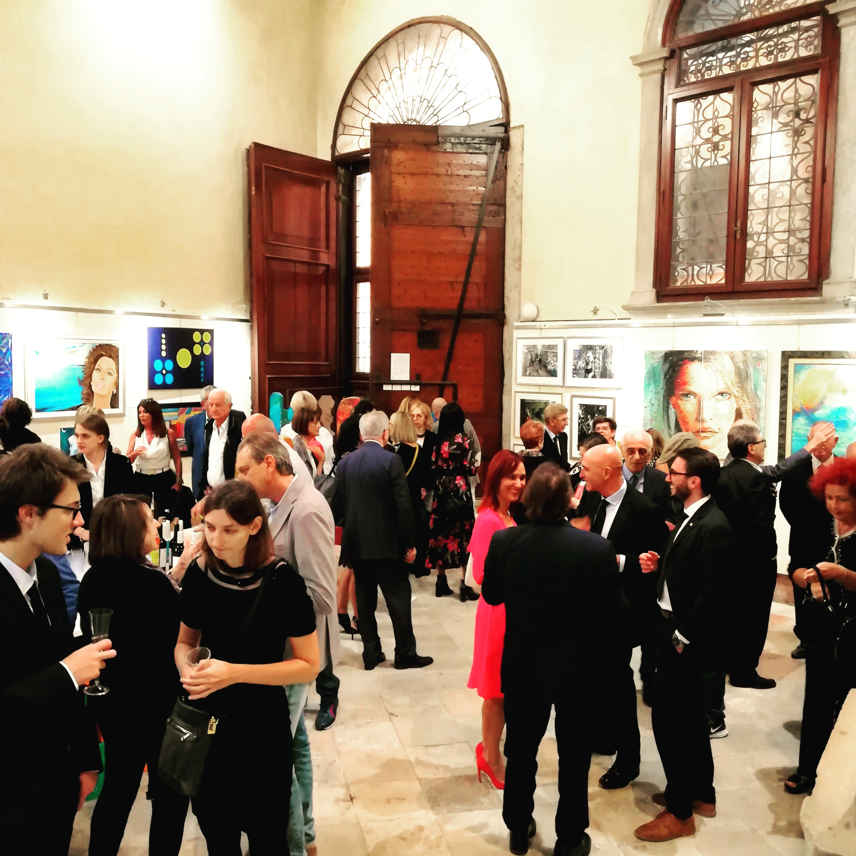 A crowded indoor gallery gathering with people standing and talking around displayed artwork, photographed in bright interior lighting.